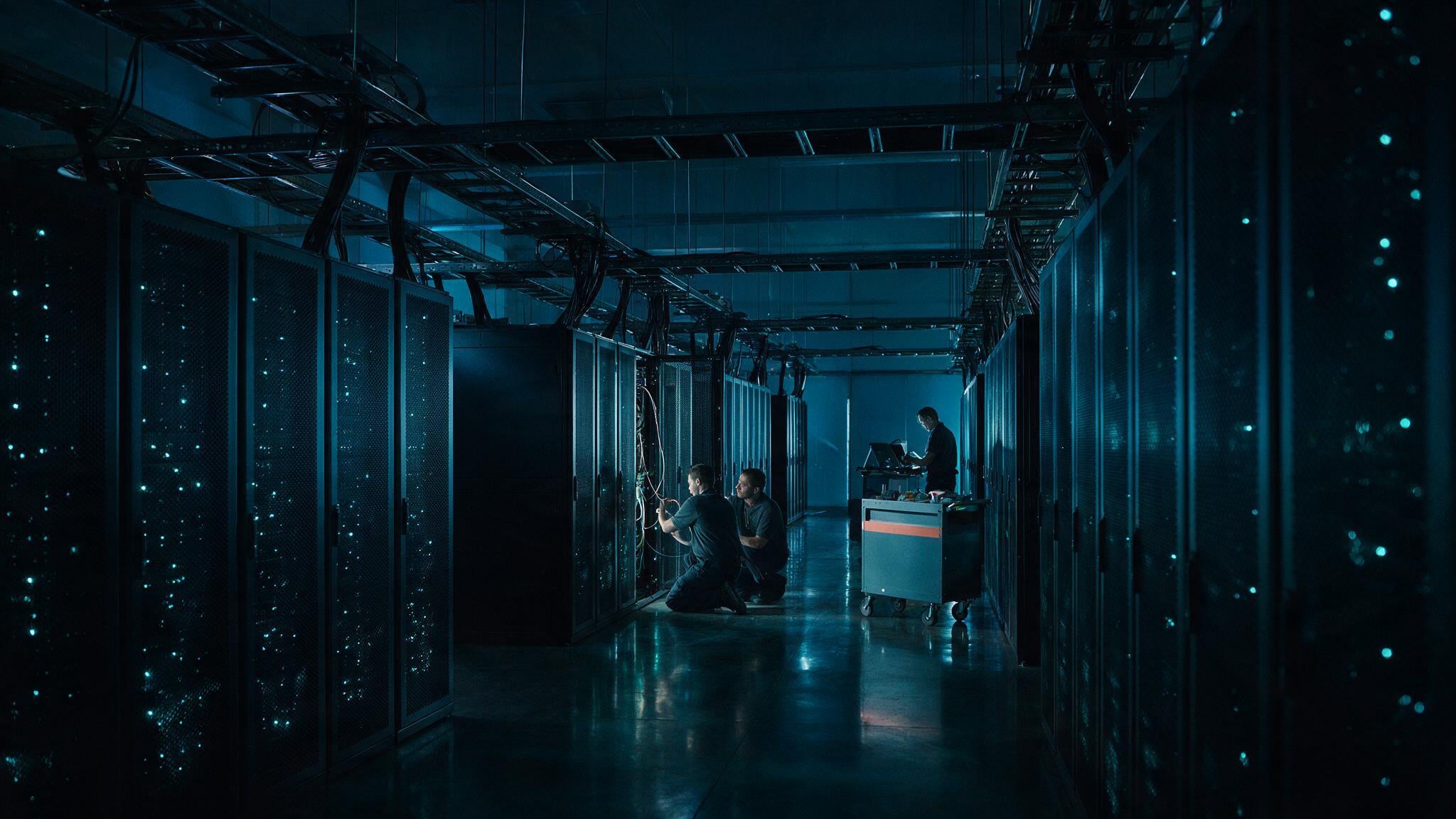 A two network engineers crouched in a cold aisle patching a dense fibre trunk into a switch chassis while a third stands at a mobile console.